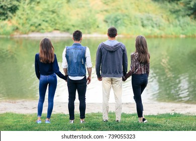 Two Love Couples Standing By Lake In Park Back View. Romantic Rest Of Four Young People In Nature, Two Girls And Two Guys Looking At Calm Water. Contemplation, Harmony, Relationship Concept