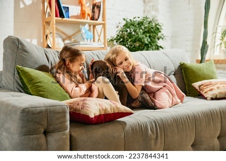 Two lovely little girls, children, sisters playing, hugging beautiful purebred dog, brown labrador at home. Love. Concept of family, childhood, pets, care, friendship, emotions and leisure