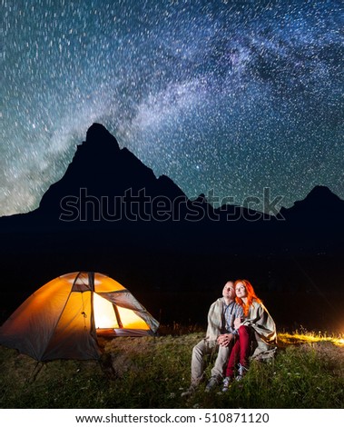 Two lovers hikers covered with a plaid sitting together near campfire and shines camp at night under stars and looking to the starry sky and Milky way. Silhouette of high mountains on background