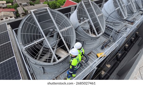 Two maintenance workers check large cooling towers on a commercial rooftop during the day.