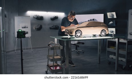 Two Male Automotive Designers Working On Model Of Eco Friendly Electric Car In Modern Car Design Studio. One Working In 3D Modeling Computer Software, Other Sculpting With Plasticine Clay And Rake.