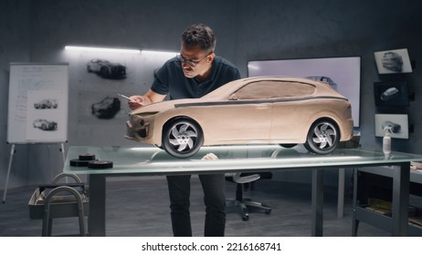 Two Male Automotive Designers Working On Model Of Eco Friendly Electric Car In Modern Car Design Studio. One Working In 3D Modeling Computer Software, Other Sculpting With Plasticine Clay And Rake.