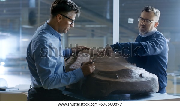 Two Male\
Automotive Designers Working on a Clay Model of New Generation\
Electric Car Future Design. One Holds Tablet Computer For Graphic\
Design, Other Sculpts with Clay with\
Rake/Wire.