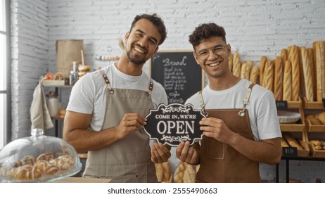 Two male bakers smiling and holding an open sign in a bakery shop filled with fresh bread loaves - Powered by Shutterstock