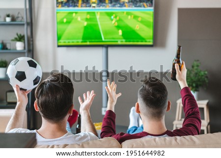 Two male buddies watching football play live broadcast on tv, cheering and drinking beer. Friends spending their day-off together at home. View from the back.