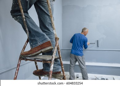 Two Male Builders Are Painting A Room. One Man Is Using A Ladder And Only His Legs Are Visible. The Other Man Is In The Background. 