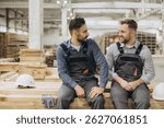 Two male construction workers in uniforms sitting on wooden planks in a factory, discussing modular building techniques and teamwork