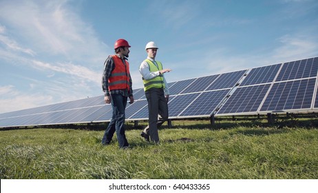 Two male electrician workers in reflective vests and hard hats walking in between long rows of photovoltaic solar panels and talking about installation of new solar panels.