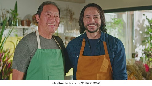 Two Male Employees Of Floriculture Store Standing Smiling. An Older And Younger Staff Of Small Business Flower Shop Looking At Camera