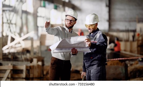 Two male engineers are standing in the building site and holding construction plan. Businessmen dressed in white protective hardhats working together and discussing detailes of design project. 