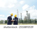 Two male engineers in uniform stand consulting work and holding tablet checking gas leakage pipe system and maintenance system and training oil refinery at column tank oil of Petrochemistry industry.