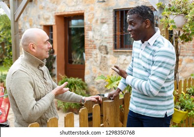 Two Male Farmers Friendly Talking Outside Next To Wooden Fence On Background With Brick House