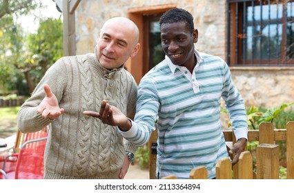 Two Male Farmers Friendly Talking Outside Next To Wooden Fence On Background With Brick House