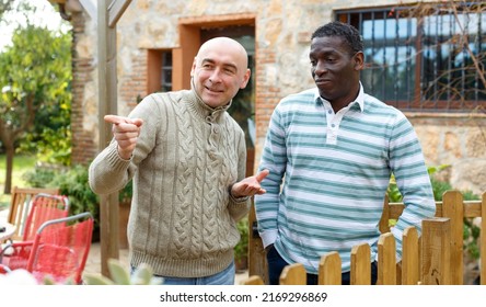 Two Male Farmers Friendly Talking Outside Next To Wooden Fence On Background With Brick House