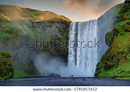 Two male and female tourists are walking at Skogafoss Falls in southern Iceland. In the morning, the sun rose from behind the mountains with green grass. This is a popular tourist destination.