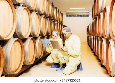 Two male and female wine factory employees wearing robes and head nets inspect oak barrel temperature controlled room maintain quality inside wine factory and record the wine fermentation clipboard. - Powered by Shutterstock