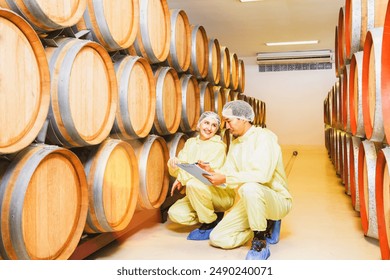 Two male and female wine factory employees wearing robes and head nets inspect oak barrel temperature controlled room maintain quality inside wine factory and record the wine fermentation clipboard. - Powered by Shutterstock