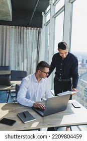 Two Male Focused Colleagues Discussing Business Project Looking At Laptop Screen During Meeting In Modern Meeting Room With Glass Walls