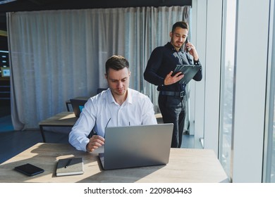 Two Male Focused Colleagues Discussing Business Project Looking At Laptop Screen During Meeting In Modern Meeting Room With Glass Walls