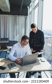 Two Male Focused Colleagues Discussing Business Project Looking At Laptop Screen During Meeting In Modern Meeting Room With Glass Walls