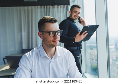 Two Male Focused Colleagues Discussing Business Project Looking At Laptop Screen During Meeting In Modern Meeting Room With Glass Walls