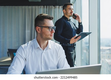 Two Male Focused Colleagues Discussing Business Project Looking At Laptop Screen During Meeting In Modern Meeting Room With Glass Walls