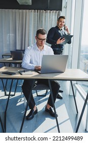 Two Male Focused Colleagues Discussing Business Project Looking At Laptop Screen During Meeting In Modern Meeting Room With Glass Walls