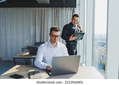 Two Male Focused Colleagues Discussing Business Project Looking At Laptop Screen During Meeting In Modern Meeting Room With Glass Walls