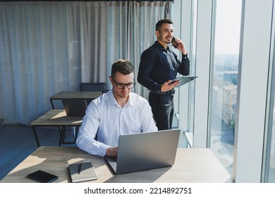 Two Male Focused Colleagues Discussing Business Project Looking At Laptop Screen During Meeting In Modern Meeting Room With Glass Walls