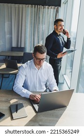 Two Male Focused Colleagues Discussing Business Project Looking At Laptop Screen During Meeting In Modern Meeting Room With Glass Walls