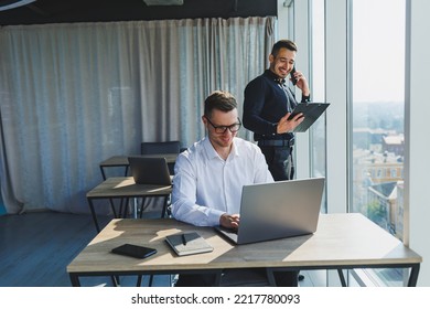 Two Male Focused Colleagues Discussing Business Project Looking At Laptop Screen During Meeting In Modern Meeting Room With Glass Walls