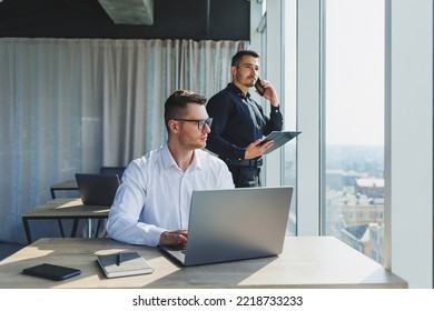 Two Male Focused Colleagues Discussing Business Project Looking At Laptop Screen During Meeting In Modern Meeting Room With Glass Walls