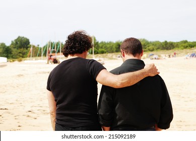 Two male friends in black clothes walking on the seaside with one of them hugging the other