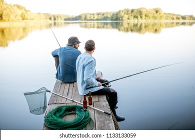 Two male friends dressed in blue shirts fishing together with net and rod sitting on the wooden pier during the morning light on the lake - Powered by Shutterstock