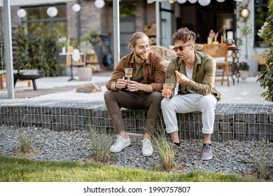 Two male friends having a close conversation, sitting with drinks on a porch of the country house outdoors. Fun summer time in a country house, male friendship