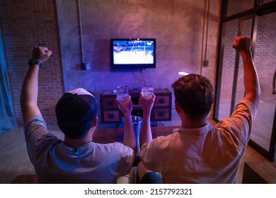 Two Male Friends Holding A Beer Glass Celebrate Watching Sports On Television At Home.