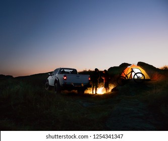 Two Male Friends Near Bonfire, Pickup Offroad Truck, Illuminated Tent And Bike At Night Camp In The Mountains. Bicycle Adventure And Car Travel Concept.