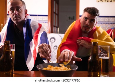 Two Male Friends And Sports Fans Are Watching A Sporting Event On Television. They Are Taking Chips From A Bowl. Each Of The Men Is Carrying A Flag On His Shoulders. Sports Concept