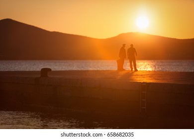 Two Male Friends Standing And Talking On The Stone Jetty During Sunset In Zedar Old Town, Croatia.