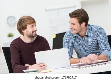 Two Male Friends Working Together In The Office Sitting Smiling And Chatting As They Share A Laptop Computer