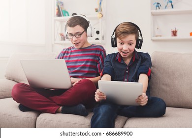 Two Male Kids With Gadgets And Headphones. Teenage Friends Playing Games On Laptop And Digital Tablet With Excitement At Home. Boy Friendship And Online Gaming Concept