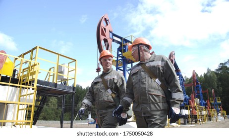 Two Male Oil Workers Walking And Talking Near Oil Pump Jacks. Engineers Overseeing Drilling Rig Of Crude Oil Production. Pumping Crude Oil For Fossil Fuel Energy With Drilling Rig In Oilfield.