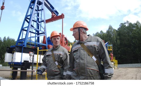 Two Male Oil Workers Walking And Talking Near Oil Pump Jacks. Engineers Overseeing Drilling Rig Of Crude Oil Production. Pumping Crude Oil For Fossil Fuel Energy With Drilling Rig In Oilfield.