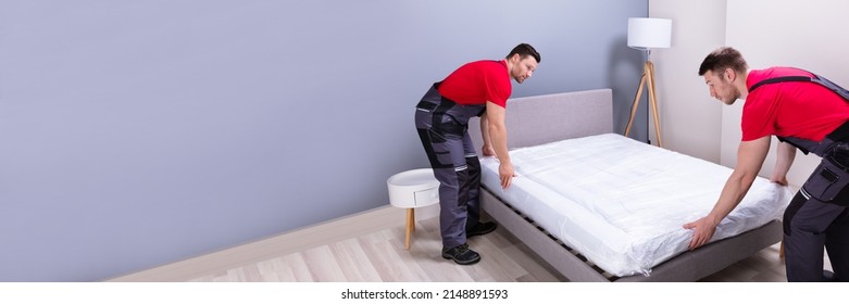 Two Male Professional Movers In Uniform Placing The Mattress Over The Bed In New House