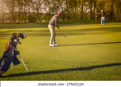 Two Male Senior Golf Player Putting On Green At Twilight, With Golf Bag In Foreground.