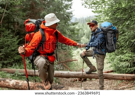 two male tourists with backpacks and hiking equipment walk in the forest together and give helping hand, people on mountain hike with trekking poles