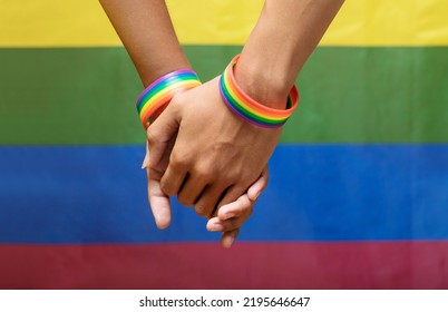Two man holding hands with a rainbow-patterned wristband on their wrists with the LGBTQ rainbow flag in the background. lgbt, same-sex relationships and homosexual concept