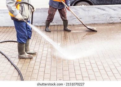 Two Man, worker in yellow orange vest and rubber boots washing sidewalk, road. Cleaning street with hose with water and shovel