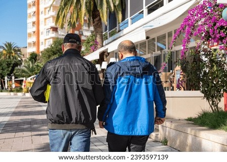 Two mans in jackets walks and talks on a city street. Male's conversation. Adult friends talking. Discussion. Senior business professionals walking together on a sunny day-Saranda Albania-Nov 23,2023