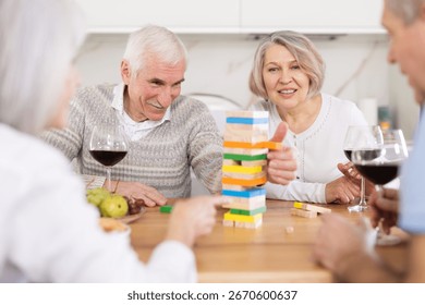 Two married couples are having fun in cozy home atmosphere, playing building tower with wooden blocks game. Senior participant is removing wooden block bricks from tower and others are watching